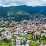 Aerial view of Budhanilkantha valley showing residential houses, green hills, and Kathmandu city skyline.