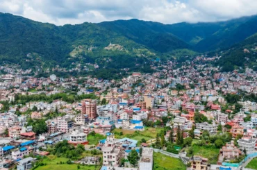 Aerial view of Budhanilkantha valley showing residential houses, green hills, and Kathmandu city skyline.