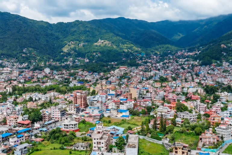 Aerial view of Budhanilkantha valley showing residential houses, green hills, and Kathmandu city skyline.